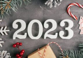 Happy New Year 2023. White wooden numbers 2023 lying on gray uneven concrete surface with Christmas tree branches and decorations, lollipop and snowflake. Merry Christmas. Top view